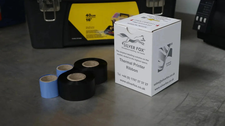 Silver Fox thermal printer ribbon box with blue and black ribbon rolls on a workbench, label printer behind.