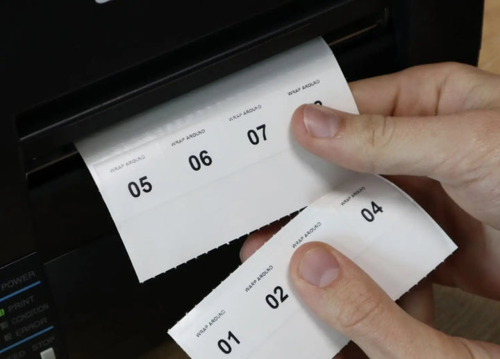 Hands pull a strip of numbered wrap-around labels from a label printer.