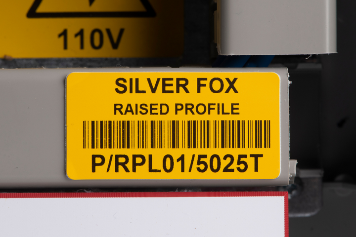 Close up of a yellow industrial identification label on equipment reading “SILVER FOX” and “RAISED PROFILE,” with a barcode and the code “P/RPL01/5025T,” with a blurred “110V” warning sign visible in the background.