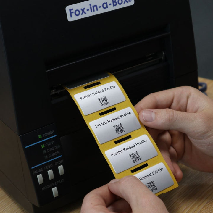 Alt text: A person feeds a strip of yellow-backed, silver “Prolab Raised Profile” labels with small QR codes out of a black “Fox-in-a-Box” label printer, with the printer’s status lights and buttons visible on the front panel.