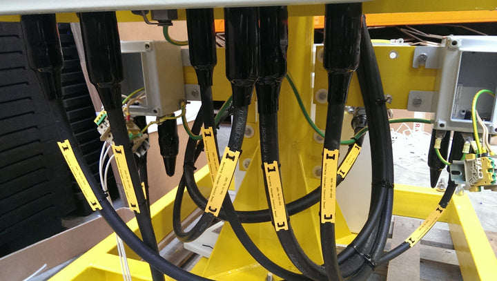 Thick black power cables inside a yellow electrical frame are tagged with yellow tie-on identification labels, with junction boxes and green earth wires nearby.