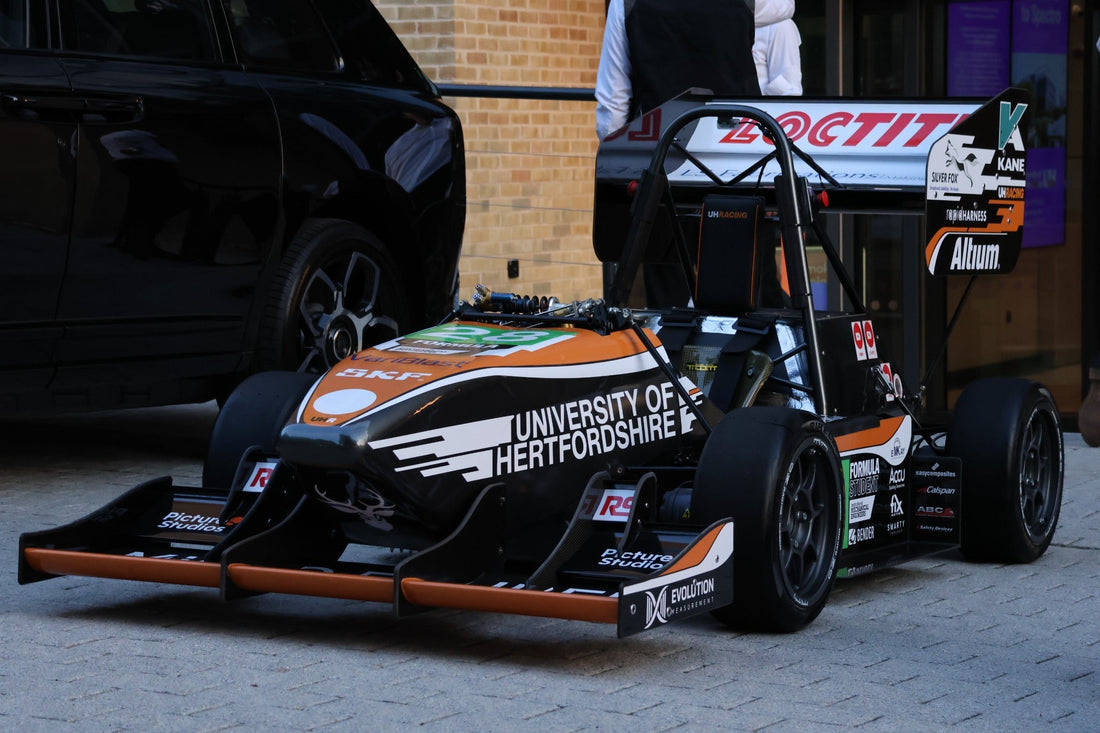 A University of Hertfordshire UH Racing single-seat formula-style race car in black, orange, and white livery is parked outdoors, with sponsor logos on the body and rear wing and a black vehicle and brick building in the background.