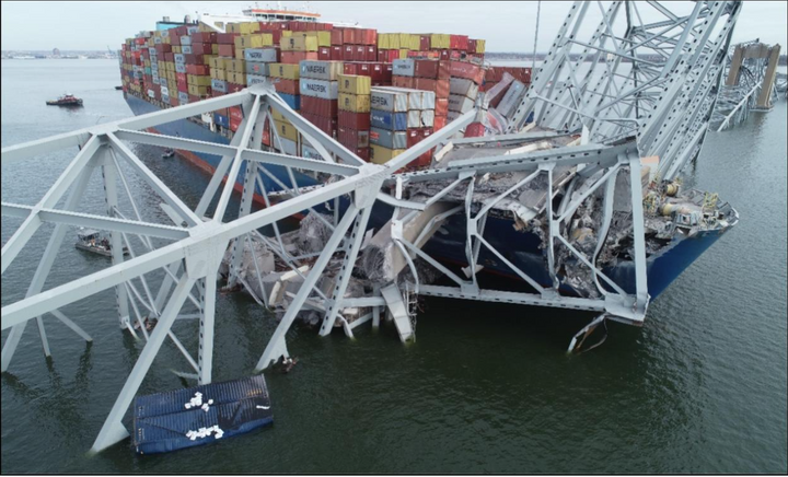 Cargo ship wedged under a collapsed steel bridge, containers stacked on deck.