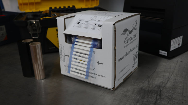 Silver Fox branded “Fox-in-a-Box” carton on a workbench, showing yellow label strips through a clear window beside a label printer.