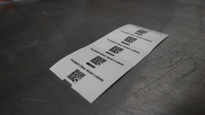 A strip of clear asset labels lies on a metal work surface, each label printed with “Prolab® Clear Asset Labels” and a QR code.