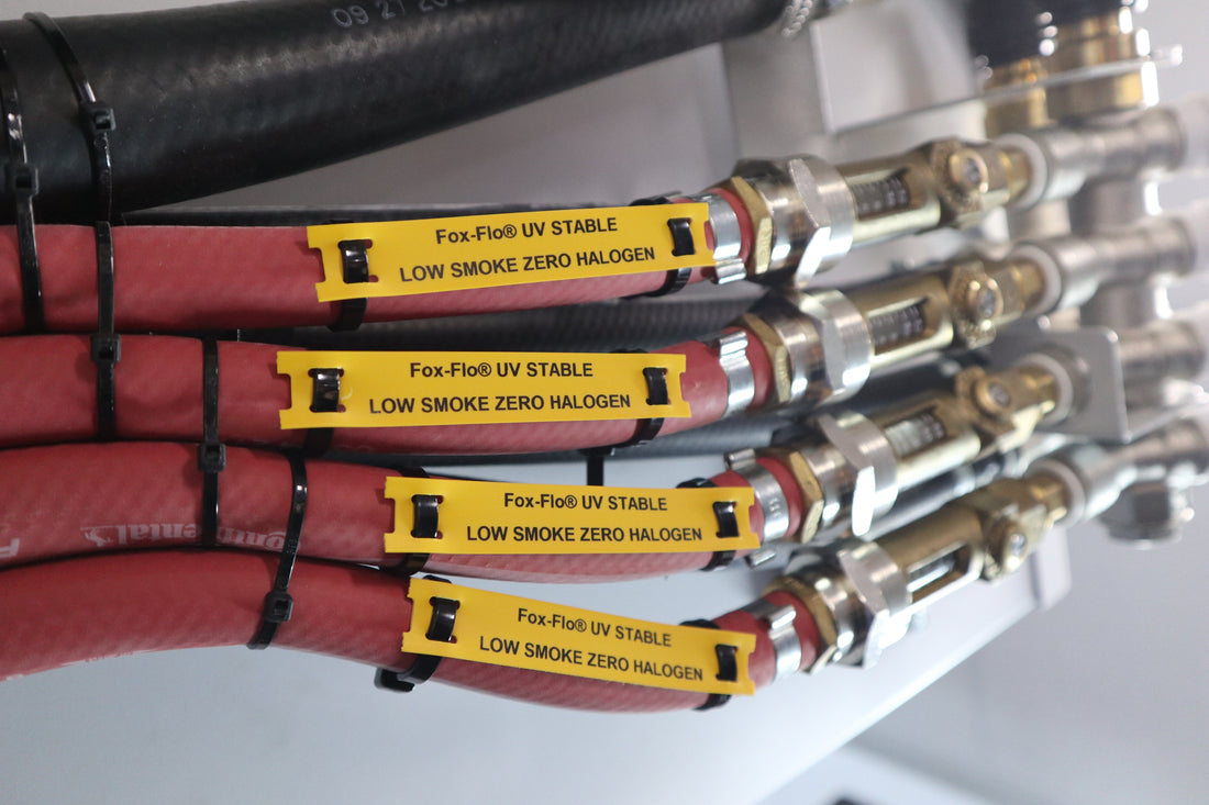 Four yellow Fox-Flo® UV Stable “Low Smoke Zero Halogen” tie-on labels are attached with black clips to red hoses near metal fittings, with additional black hoses and connectors in the background.
