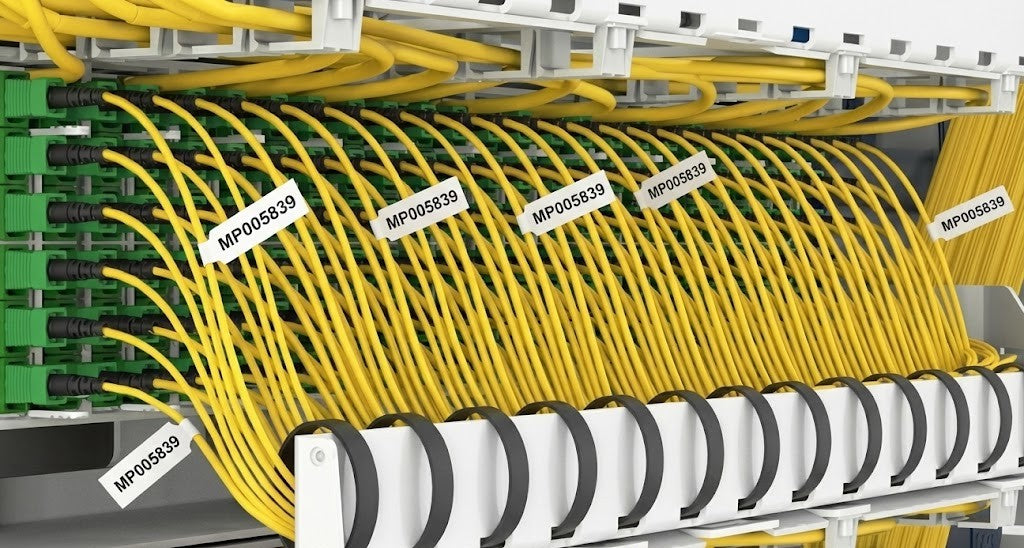 A fibre distribution panel filled with neatly routed yellow patch cords plugged into green connectors, with several small white cable labels reading “MP005839” attached along the bundle.