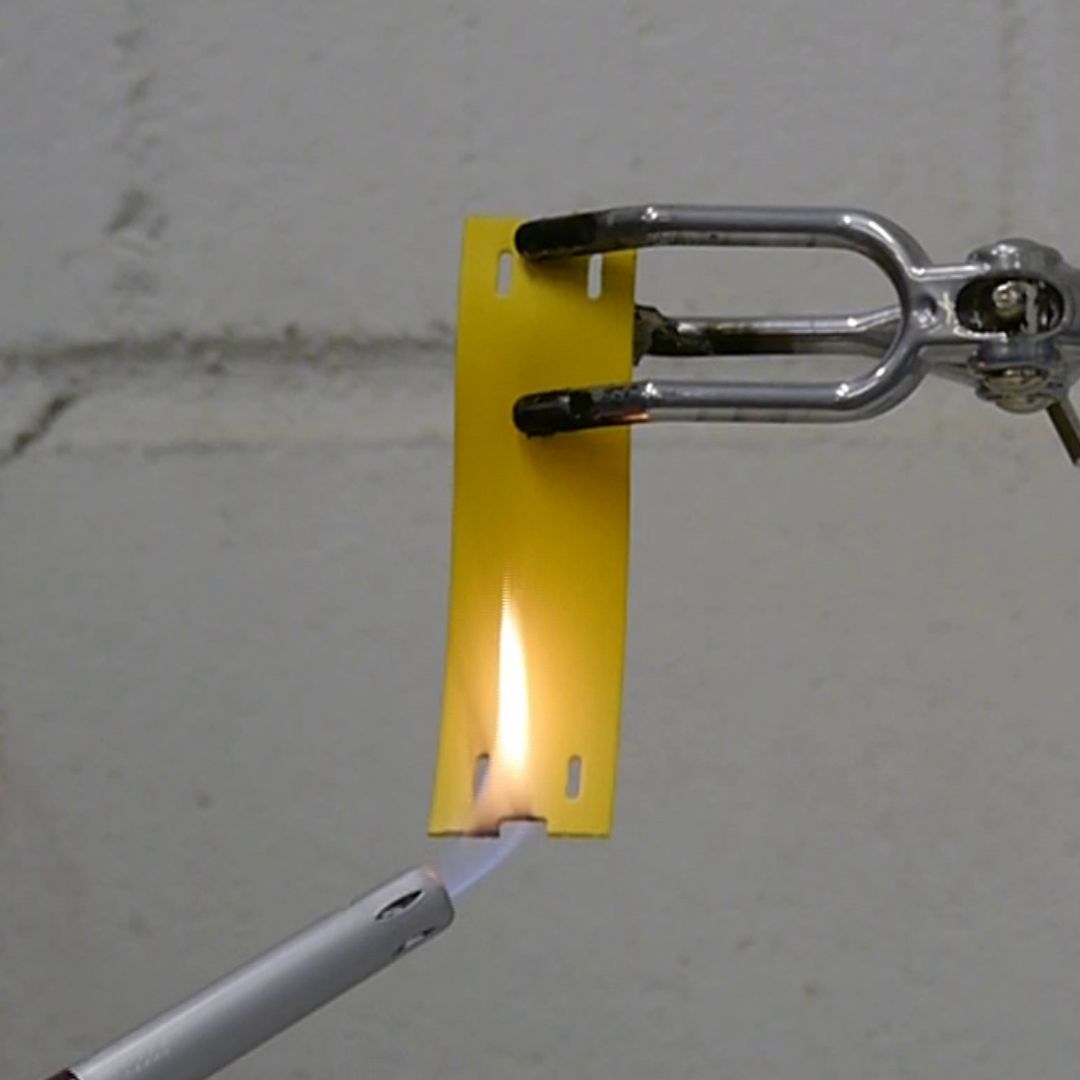 A yellow low smoke zero halogen label is clamped at the top with metal tongs while a lighter flame burns the bottom edge, showing how the material reacts to fire against a plain grey background.