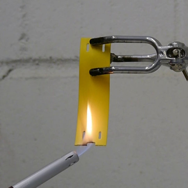 A yellow low smoke zero halogen label is clamped at the top with metal tongs while a lighter flame burns the bottom edge, showing how the material reacts to fire against a plain grey background.