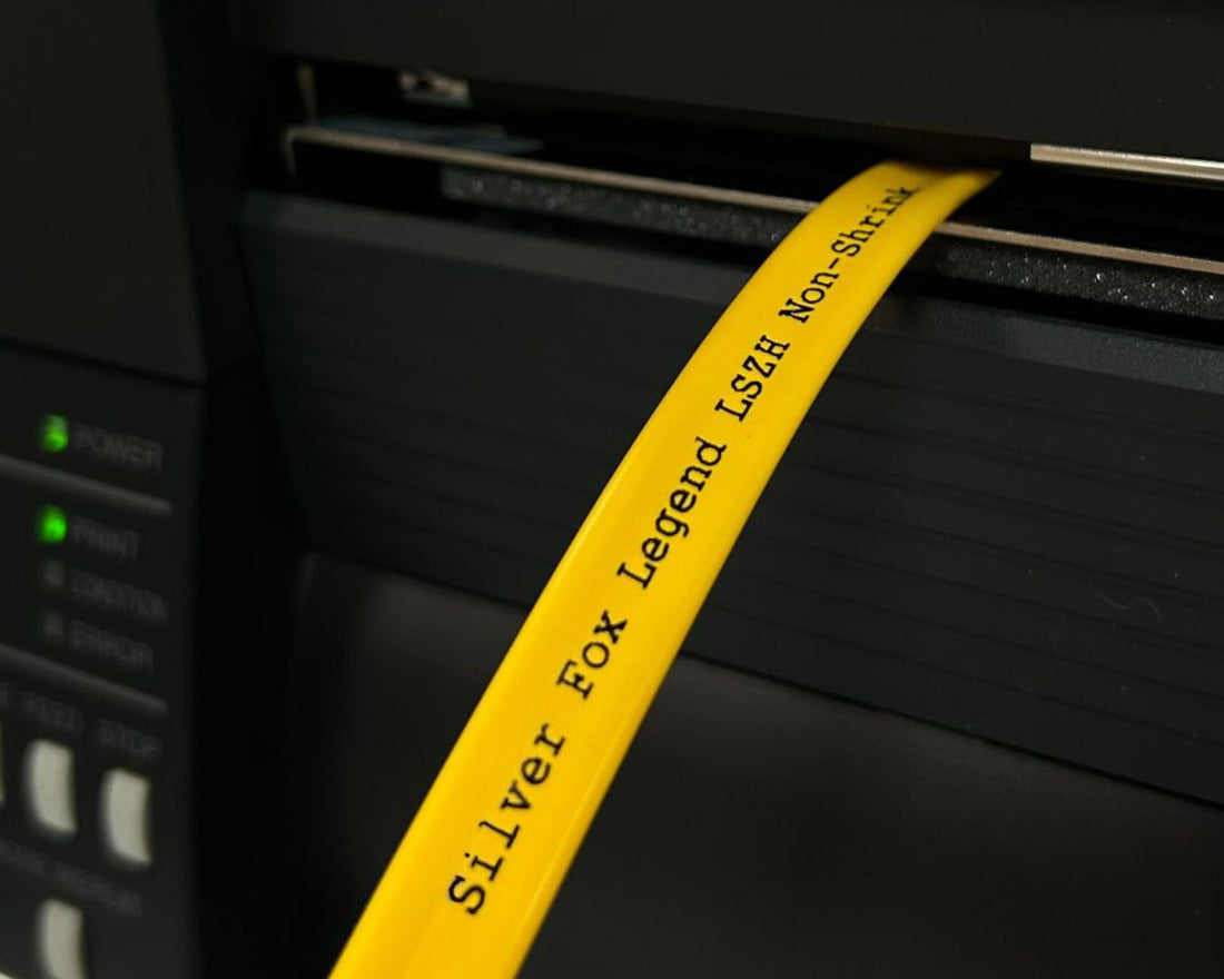 A yellow label tape printed with “Silver Fox Legend LSOH Non-Shrink” feeds out of a black label printer, with green status lights visible on the control panel.