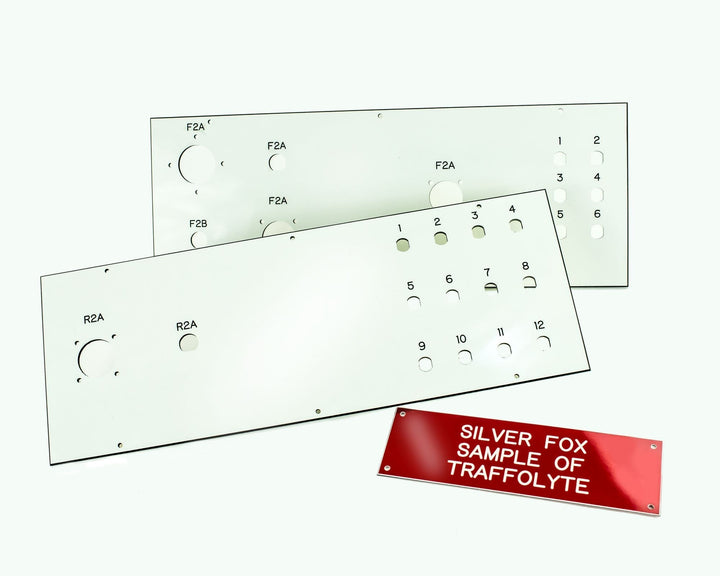 Two white Traffolyte engraved control panels with circular and square cutouts and labels (F2A, F2B, R2A, and numbered 1 to 12), laid on a white background beside a red sample plate reading “SILVER FOX SAMPLE OF TRAFFOLYTE.”