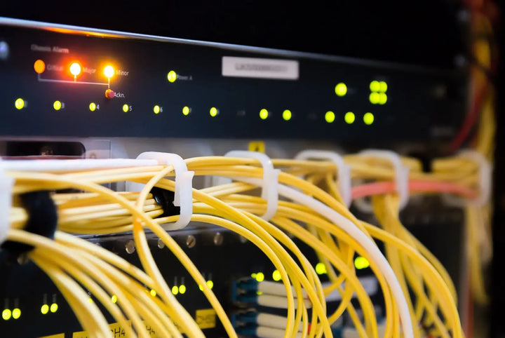 Close-up of a network rack with bundles of yellow fiber optic cables secured by white clips in the foreground, and a router or switch panel behind them with glowing green and amber status lights.