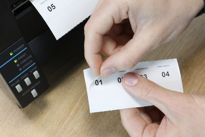 Hands peel a numbered white wrap-around label strip (01–04) on a wooden desk next to a label printer, with another printed strip showing “05” emerging from the printer.