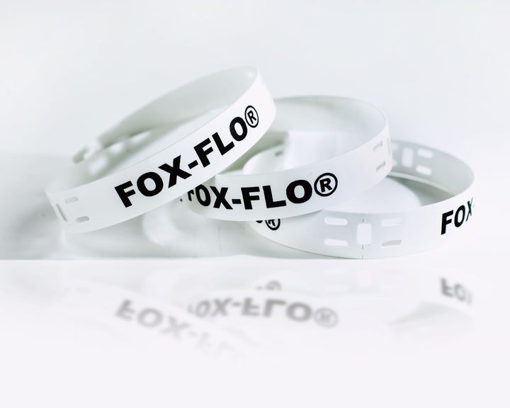 Coiled white cable marker strips printed with “FOX-FLO®” in bold black lettering lie on a clean white surface, with the slotted ends visible for fastening.