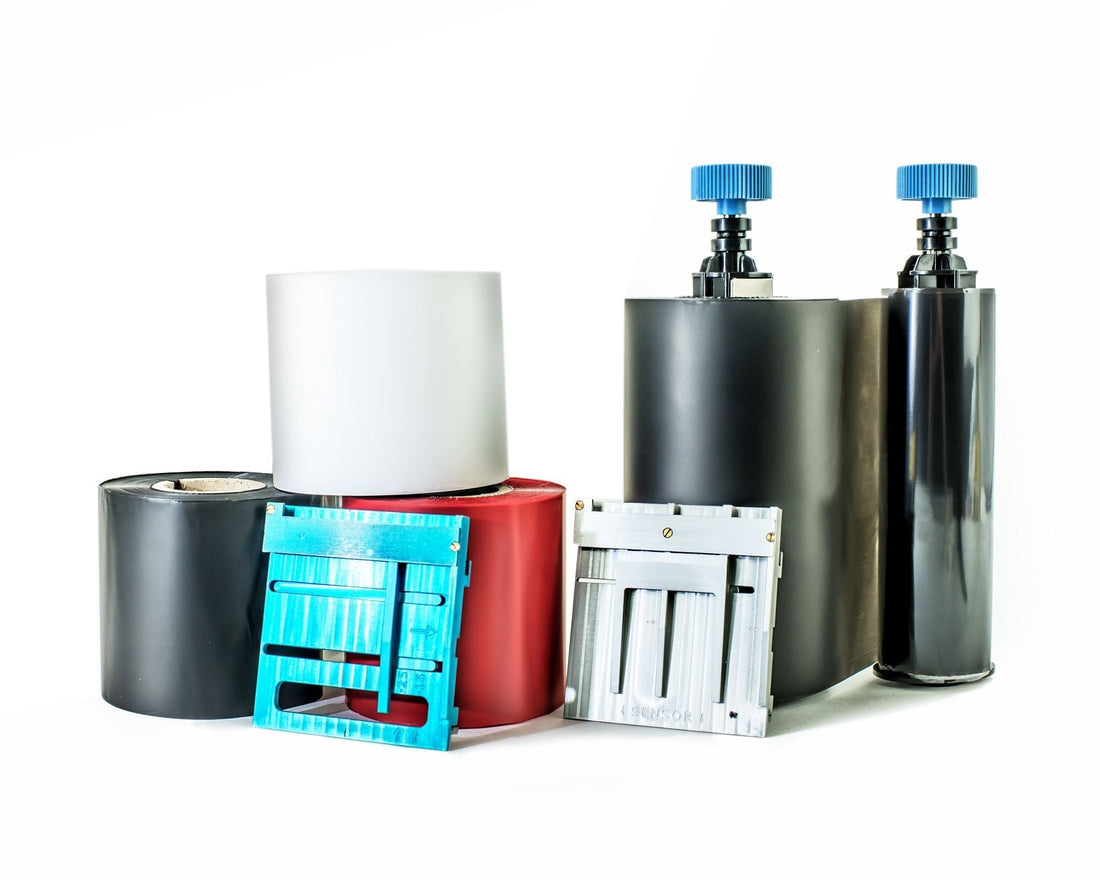Assorted thermal transfer printer ribbons, cartridges, and two black bottles with blue caps on a white background.