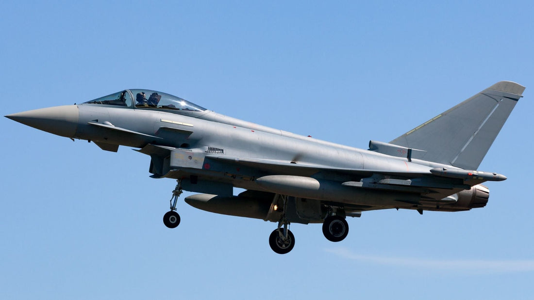 A grey fighter jet flies against a clear blue sky with its landing gear down, viewed from the side with the cockpit and pilot visible.